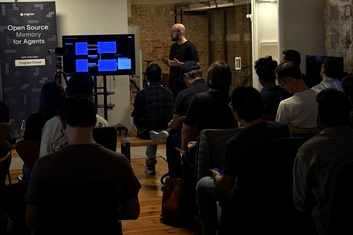A speaker stands beside a screen showing a blue architecture diagram, next to a cognee roll-up banner reading Open Source Memory for Agents and Now available: Cognee Cloud. A seated audience fills the dim foreground of a loft room.